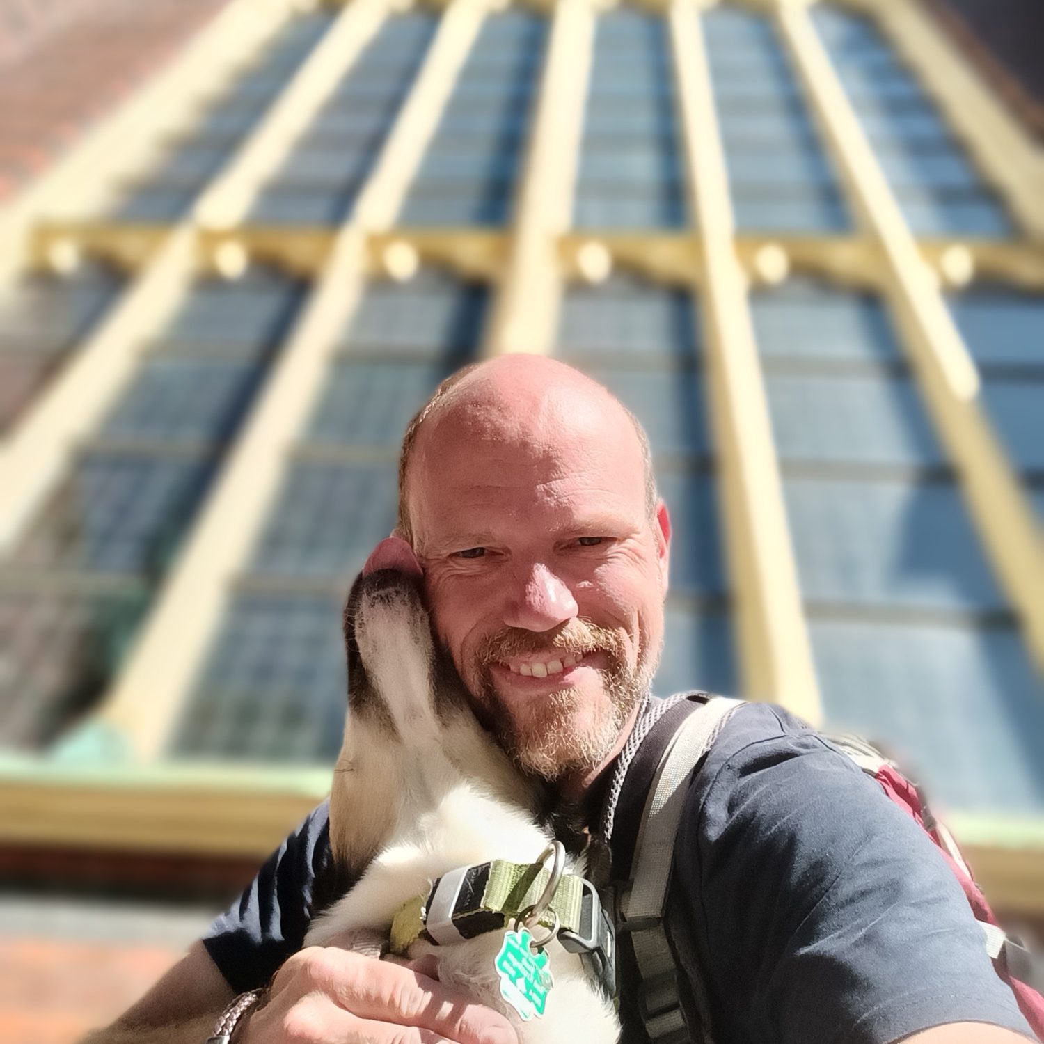 Chris mit Hund Sammy, der ihm einen Kuss gibt, vor einer großen Fensterglaskonstruktion.