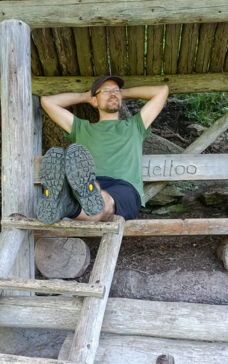 Chris sitzt entspannt unter einem Holzdach mit hochgelegten Füßen.