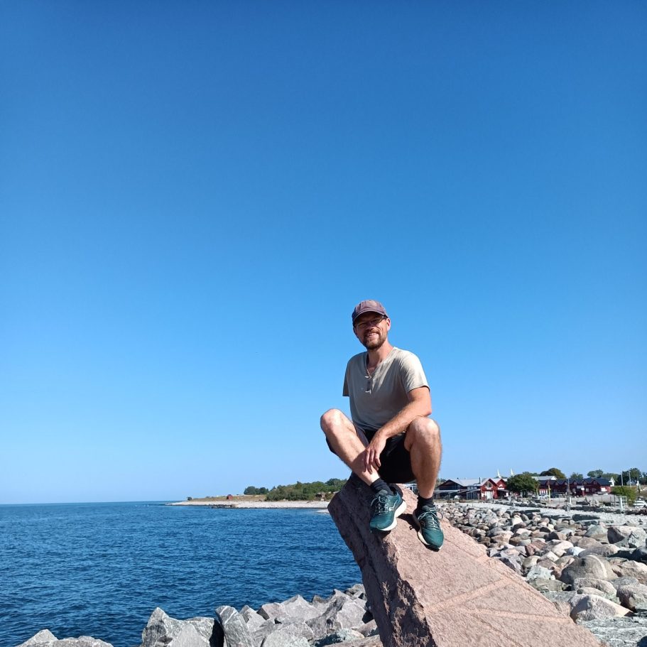 Chris sitzt auf Felsen am Ufer, blauer Himmel und ruhige See im Hintergrund.